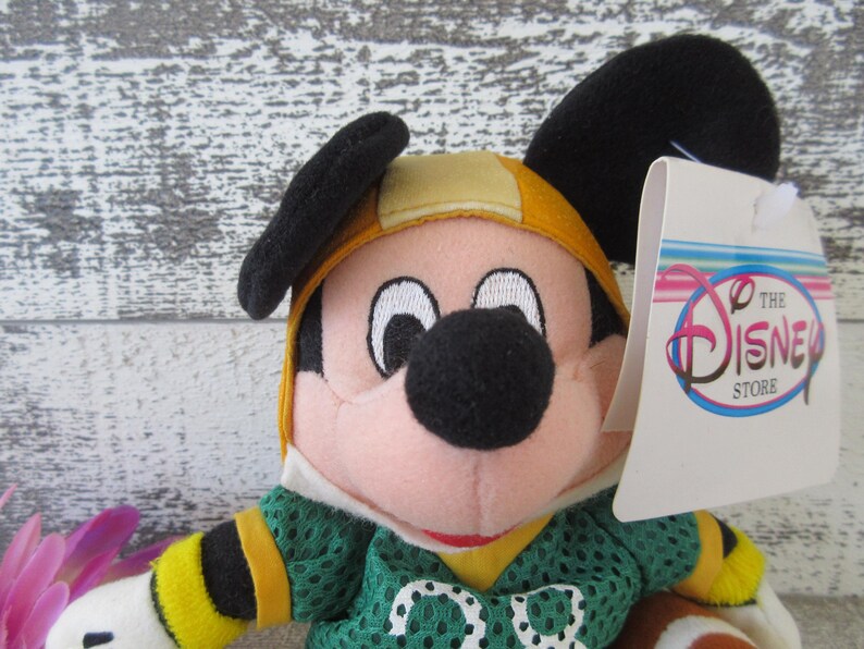 May include: A Mickey Mouse plush toy wearing a green football jersey with the number 00. The toy has a yellow helmet and black ears. A tag from The Disney Store is attached.