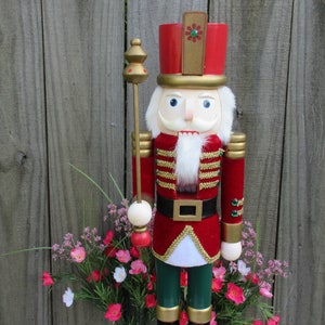 Könnte beinhalten: Eine traditionelle Holz-Nussknacker-Figur mit einer rot-goldenen Uniform, einem weißen Bart und einem grünen Hut. Der Nussknacker hält einen goldenen und roten Stab.