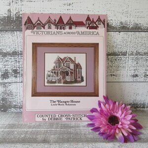 May include: A cross-stitch pattern titled "The Hanger House" from the "Victorians Across America" series. The pattern features a detailed illustration of a Victorian house. A pink and purple flower rests beside the pattern. The text "COUNTED CROSS-STITCH by DEBBIE PATRICK" is visible.