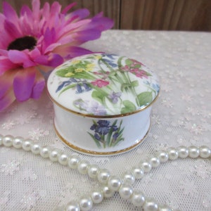 May include: A white porcelain trinket box with a floral design featuring bluebells, primroses, and other wildflowers. The box has a gold trim around the lid and base.