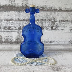 May include: A cobalt blue glass bottle shaped like a violin, with a cork stopper. The bottle sits on a light blue doily, with a strand of white pearls in front. The background is a distressed white and gray wooden surface.