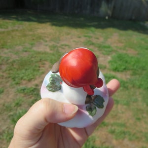 May include: A small porcelain figurine of a girl wearing a red hat and sitting on a white base with green leaves.