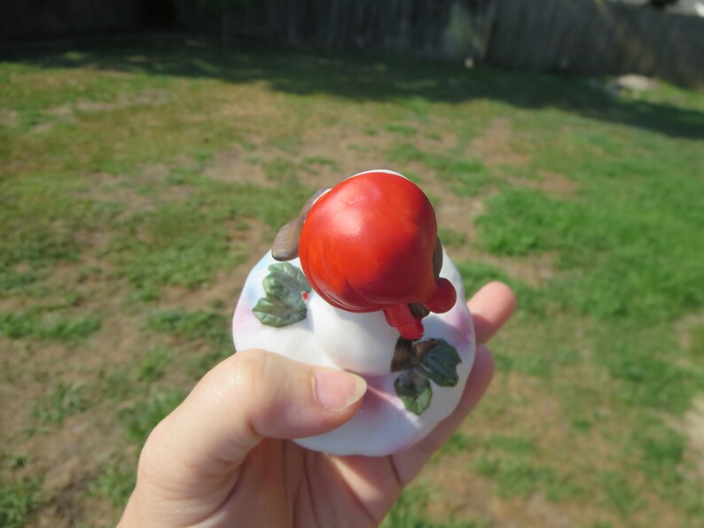 May include: A small porcelain figurine of a girl wearing a red hat and sitting on a white base with green leaves.