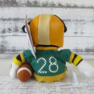 May include: A plush toy in a football uniform, viewed from the back. The toy has a yellow helmet with a tan stripe, a green jersey with the number 28, and a brown football. The arms have black and yellow stripes.