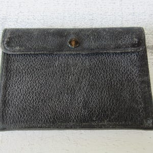 May include: A rectangular, black leather wallet with a textured surface. The wallet has a flap closure secured by a small, round button. The leather appears worn, suggesting age and use. The wallet is closed.