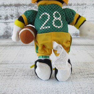 May include: A plush toy dressed as a football player, wearing a green jersey with the number 28 in white. The toy is holding a brown football and is wearing yellow pants and black and white shoes.