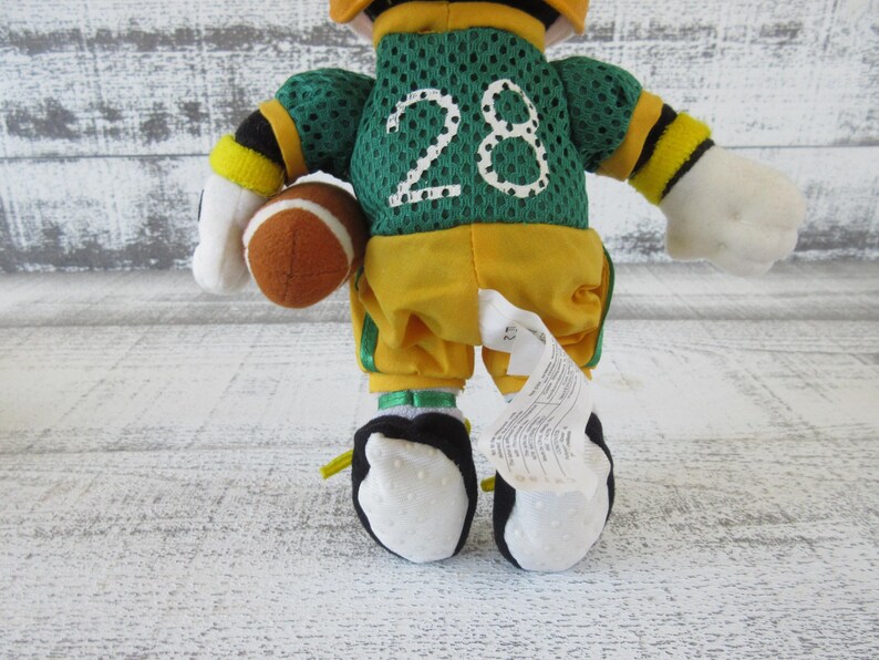 May include: A plush toy dressed as a football player, wearing a green jersey with the number 28 in white. The toy is holding a brown football and is wearing yellow pants and black and white shoes.