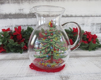 May include: A clear glass pitcher with a Christmas tree design. The tree is decorated with ornaments, lights, and a star on top. The pitcher is sitting on a red crochet doily.