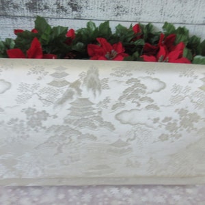 May include: A white satin clutch purse with an embossed design of a Japanese garden scene. The design features trees, pagodas, and clouds.