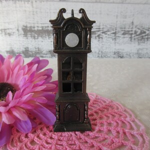 Puede incluir: Un sacapuntas de madera marr&oacute;n con forma de reloj de pie. La esfera del reloj tiene un c&iacute;rculo blanco en el centro. El sacapuntas est&aacute; sobre un encaje de ganchillo rosa.