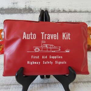 May include: Red vinyl auto travel kit with white lettering that reads "Auto Travel Kit", "First Aid Supplies", and "Highway Safety Signals". A small black and white illustration of a car with an "SOS" flag is above the text.