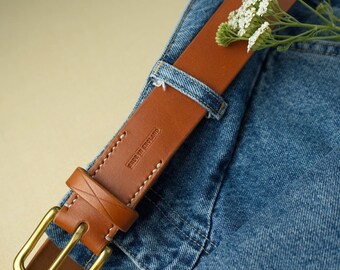 Puede incluir: Un cinturón de cuero marrón con una hebilla dorada y el texto "Made in England" en la correa. El cinturón está sobre un par de pantalones vaqueros azules.