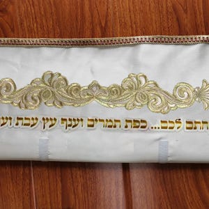 Puede incluir: Un artículo de tela blanca con bordados y texto dorados. El bordado dorado presenta un diseño floral decorativo en el centro. Los bordes están adornados con un ribete decorativo dorado y marrón. El texto es dorado.