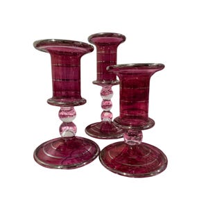 May include: Three magenta glass candle holders of varying heights, each featuring a wide circular base, a central stem with two clear glass spheres, and a cylindrical top section decorated with thin, horizontal silver-toned metallic stripes.