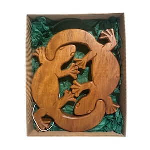 Puede incluir: Una talla de madera marrón que presenta dos figuras de lagarto entrelazadas dispuestas en un patrón circular. La madera lisa y pulida muestra una textura de grano natural. La talla se coloca dentro de una caja de cartón rectangular forrada con papel de seda verde.