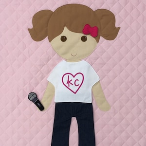 May include: A felt doll with brown hair, a white shirt with a pink heart and the letters "KC" inside, blue jeans, and a microphone. The doll is standing on a pink quilted background.