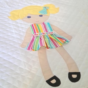 May include: A colorful fabric doll with a rainbow striped dress and a birthday hat. The dress has a white ribbon with the text "Happy Birthday" on it. The doll has blonde hair and black shoes.