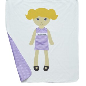 May include: A white and purple paper doll blanket with a girl doll on the front. The doll has blonde hair, a purple dress with white polka dots, and black shoes. The text "Paper Doll Blanket" is printed on the doll's dress.