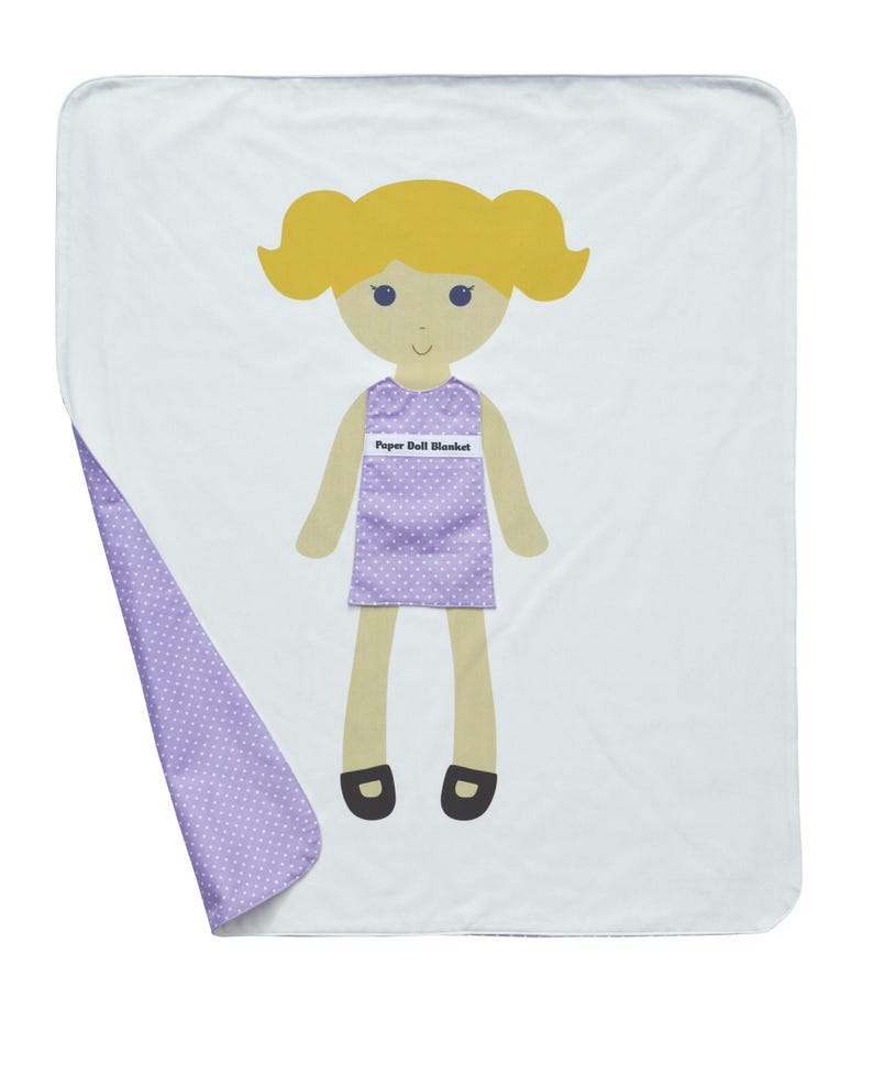 May include: A white and purple paper doll blanket with a girl doll on the front. The doll has blonde hair, a purple dress with white polka dots, and black shoes. The text "Paper Doll Blanket" is printed on the doll's dress.