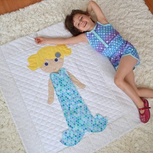 May include: A white quilted blanket with a mermaid applique. The mermaid is wearing a blue and green patterned tail and has blonde hair and a blue bow. A young girl is lying on the blanket.