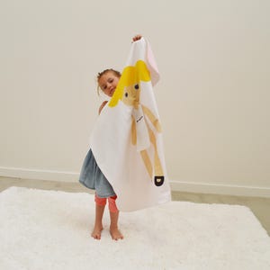 May include: A young person holding a white blanket with a yellow doll printed on it. The doll has blonde hair and is wearing a white dress. The blanket has a pink border.