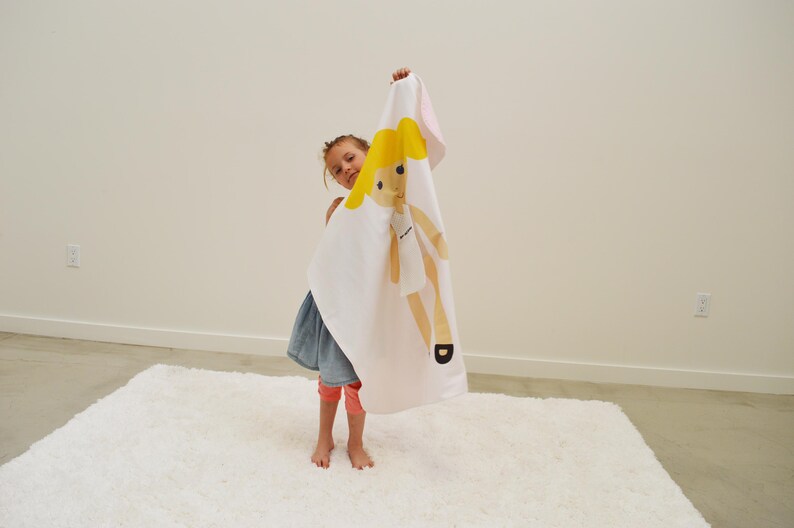 May include: A young person holding a white blanket with a yellow doll printed on it. The doll has blonde hair and is wearing a white dress. The blanket has a pink border.
