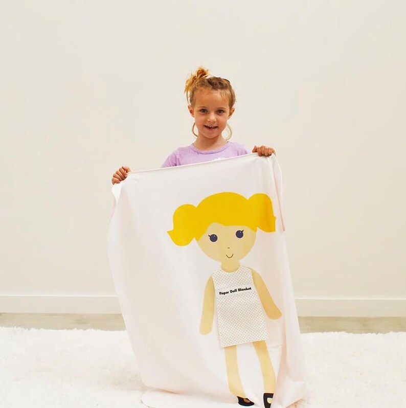 May include: A white blanket with a paper doll design. The doll has yellow hair, a white dress, and the text "Paper Doll Blanket" printed on the dress.