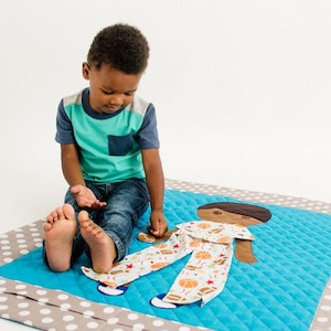 May include: A child plays with a blue and white quilted playmat featuring a brown and white appliqué of a person. The playmat has white polka dots on a gray background.