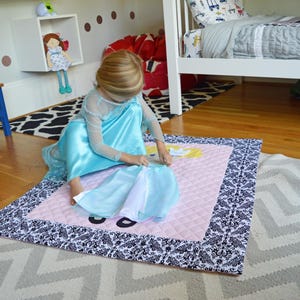Könnte beinhalten: Ein junges Mädchen in einem hellblauen Kleid spielt mit einer rosa und weißen gesteppten Spielmatte mit einem schwarzen und weißen Rand. Die Matte hat ein Prinzessinnen-Design.