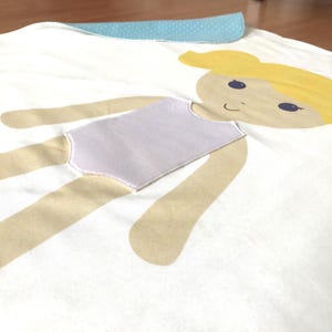 May include: A white fabric doll with a light brown body, a purple swimsuit, and a yellow head with a blue face and brown eyes.