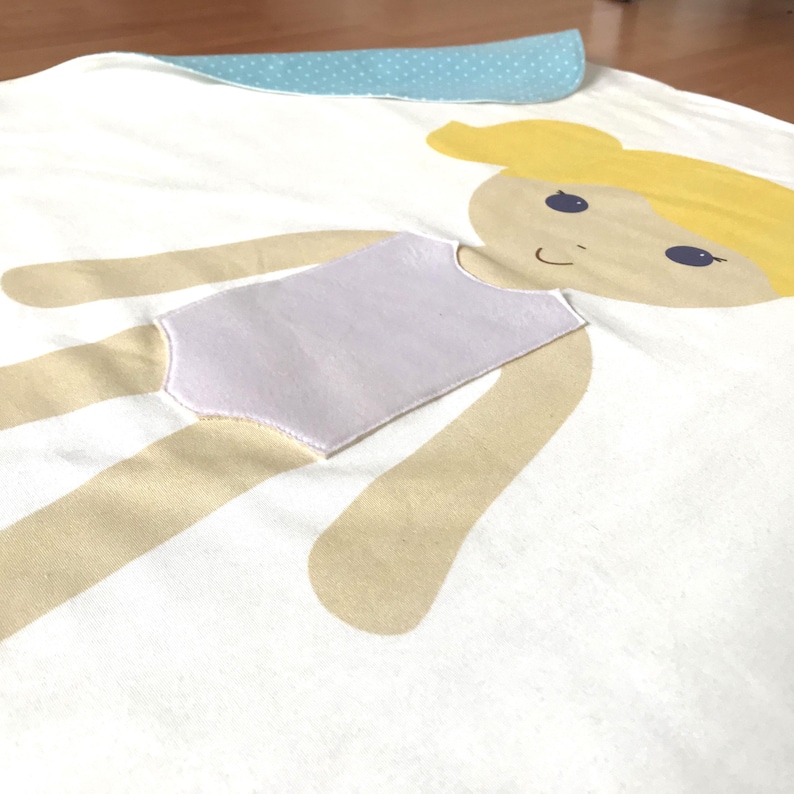 May include: A white fabric doll with a light brown body, a purple swimsuit, and a yellow head with a blue face and brown eyes.