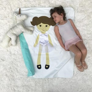 May include: A white blanket with a green and white border. A girl doll with brown hair and a white dress is sewn onto the blanket. A white unicorn stuffed animal is on the blanket. A young girl is lying on the blanket.