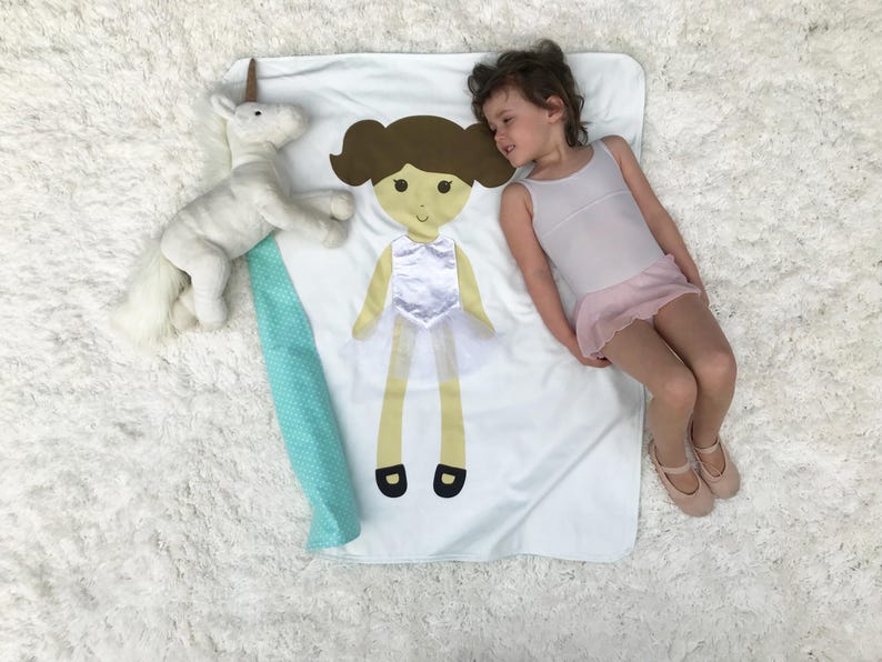 May include: A white blanket with a green and white border. A girl doll with brown hair and a white dress is sewn onto the blanket. A white unicorn stuffed animal is on the blanket. A young girl is lying on the blanket.