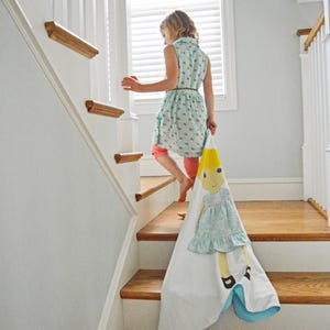May include: A young person in a light blue dress with floral print walks down a wooden staircase, carrying a white blanket with a colorful doll illustration.