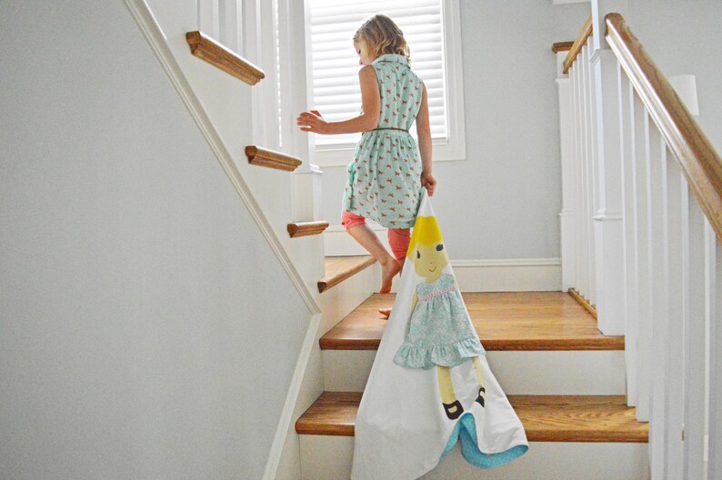 May include: A young person in a light blue dress with floral print walks down a wooden staircase, carrying a white blanket with a colorful doll illustration.