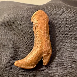 May include: A small, detailed, brown cowboy boot figurine. The boot has a pointed toe, a stacked heel, and decorative stitching. The boot is made of a textured material and appears to be a miniature decorative item.