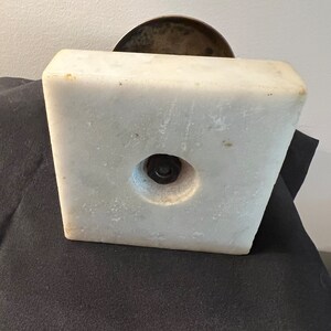 Op de afbeelding: Een witte marmeren basis met een ronde, bronzen bovenkant. De basis heeft een ronde opening in het midden met een metalen schroef.