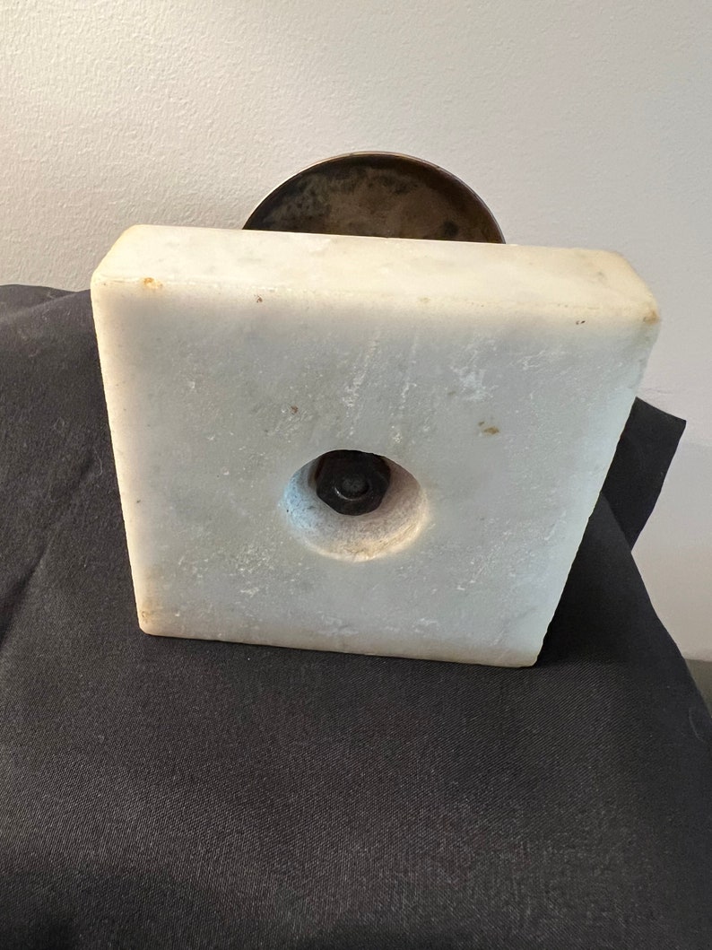 Op de afbeelding: Een witte marmeren basis met een ronde, bronzen bovenkant. De basis heeft een ronde opening in het midden met een metalen schroef.