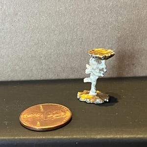 May include: A miniature gold-colored metal stand with a white cherub figure holding a round tray. The stand is about the size of a US penny.