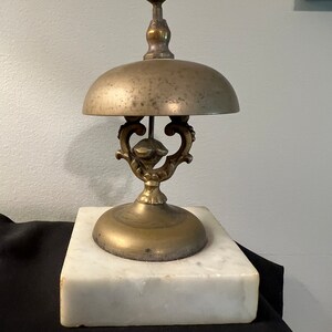 Op de afbeelding: Een vintage messingbel met een decoratieve basis en een marmeren voet. De bel heeft een verweerde afwerking en een delicaat ontwerp.