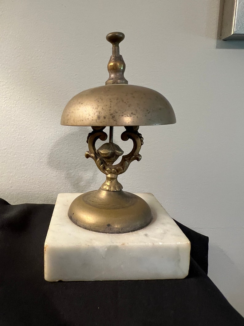 Op de afbeelding: Een vintage messingbel met een decoratieve basis en een marmeren voet. De bel heeft een verweerde afwerking en een delicaat ontwerp.