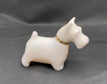 May include: White ceramic figurine of a Scottish Terrier dog wearing a gold collar. The figurine is standing on a black surface.