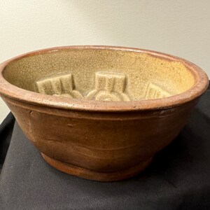 May include: A brown ceramic mold with a light brown glaze and a crackled finish. The mold has a circular shape with a raised design in the center.