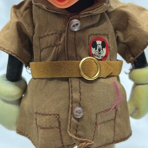 May include: Close-up of a vintage Mickey Mouse figurine wearing a tan safari-style outfit. The outfit includes a collared shirt with pockets, a belt, and a circular patch with Mickey's image and the words "Mickey Mouse".