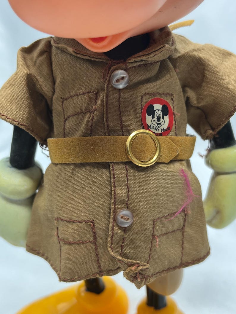 May include: Close-up of a vintage Mickey Mouse figurine wearing a tan safari-style outfit. The outfit includes a collared shirt with pockets, a belt, and a circular patch with Mickey's image and the words "Mickey Mouse".