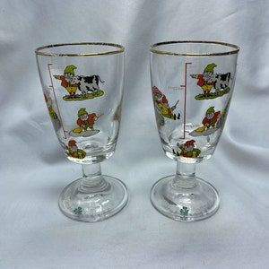 Op de afbeelding: Twee heldere glazen Irish coffee glazen met een gouden rand. Elk glas heeft een cartoon-dwergontwerp met een koe, een paddenstoel en een schop. De bodem van elk glas heeft een klavertjevier en de woorden "Irish Coffee".