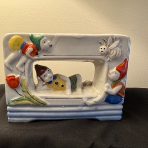 Puede incluir: Un macetero de cerámica blanca con forma rectangular y un recorte en el centro. El macetero está decorado con figuras coloridas de duendes y una mariposa. Las figuras están pintadas en tonos de rojo, amarillo, azul y verde.