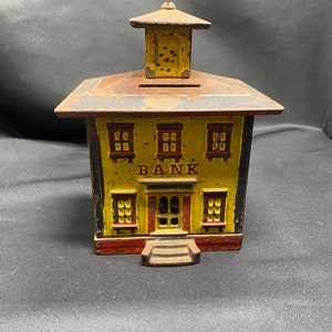 May include: A vintage cast iron bank shaped like a yellow building with a red roof and a sign that reads "BANK". The bank has three windows on each side and a door with a small set of steps.