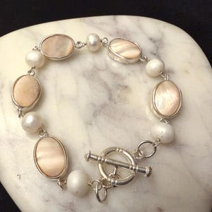 May include: A silver bracelet featuring oval, light pink mother-of-pearl stones and white pearls. The bracelet has a toggle clasp and is displayed on a white, marble-like surface. The bracelet is a piece of jewelry.