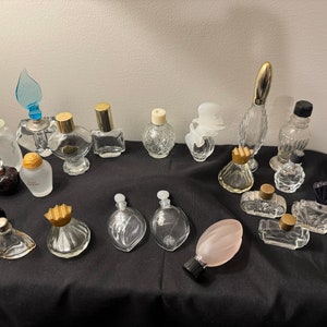 May include: A collection of vintage perfume bottles in various shapes and sizes. The bottles are made of clear glass and have gold and silver accents. Some of the bottles have decorative stoppers, including a butterfly design and a heart shape.